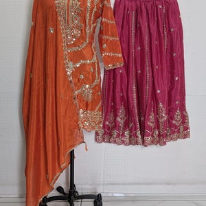 Puede incluir: Un conjunto de seda naranja con una t&uacute;nica de manga larga con intrincados bordados dorados y lentejuelas, combinada con una bufanda drapeada a juego. Tambi&eacute;n se muestra una falda rosa fucsia con bordados dorados.
