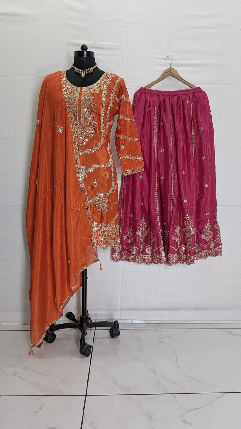 Puede incluir: Un conjunto de seda naranja con una t&uacute;nica de manga larga con intrincados bordados dorados y lentejuelas, combinada con una bufanda drapeada a juego. Tambi&eacute;n se muestra una falda rosa fucsia con bordados dorados.