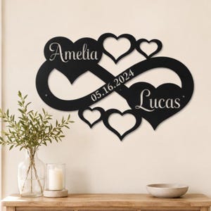 Op de afbeelding: Een zwart metalen wandbord met een oneindigheidssymbool verweven met vier hartvormen. De naam Amelia is uit het linkerhart gesneden, de naam Lucas uit het rechterhart en de datum 05.16.2024 staat in het midden van het symbool.