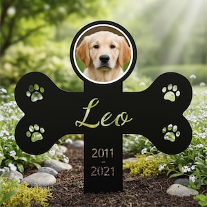 May include: A black metal pet memorial in the shape of a bone, with a circular photo of a golden retriever. The name "Leo" is inscribed in gold script, along with the years 2011-2021. Paw prints are cut out on the bone.