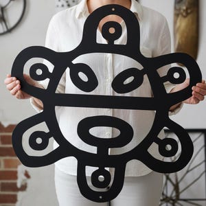 May include: A large, black metal wall art piece with a unique, abstract design. The artwork features a series of interconnected shapes, including circles and curved lines, creating a stylized, organic form. The piece is held up by a person.