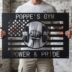 Peut inclure: Panneau métallique noir avec le texte "POPPE'S GYM POWER & PRIDE". Le panneau présente un motif découpé d'une main d'haltérophile tenant un haltère et une section remplie d'étoiles ressemblant à un drapeau.