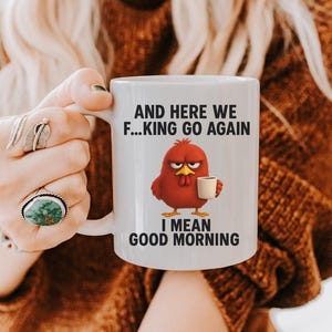 May include: White ceramic mug with the text "AND HERE WE F...KING GO AGAIN I MEAN GOOD MORNING" and a cartoon red chicken holding a coffee cup. The mug has a white handle and is held by a person.