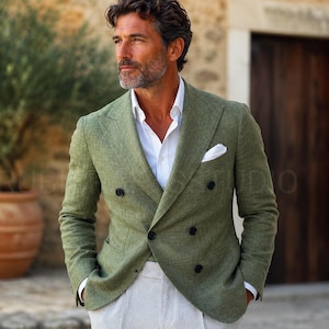 May include: A double-breasted olive green blazer with black buttons, paired with a white dress shirt and light gray pants. A white pocket square is visible in the blazer's breast pocket. The blazer is made of a textured fabric.