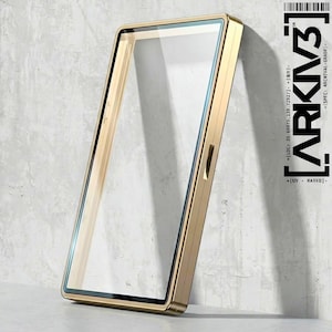 May include: A gold-colored rectangular phone case with a clear glass screen protector. The case is leaning against a textured gray wall. The text "ARKIV3" and other details are printed on the right side of the image.