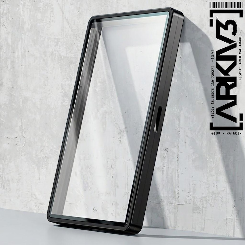 May include: A black rectangular frame with a clear glass panel, leaning against a textured gray wall. The frame has a sleek, modern design. The text "ARKIV3" and other technical details are printed on the right side of the frame.
