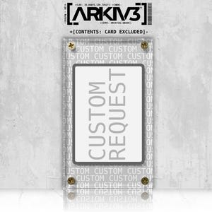 Puede incluir: Una vitrina de acrílico transparente con el texto "CUSTOM REQUEST" en gris. La caja está montada con cuatro tornillos de latón. La parte superior de la caja tiene el texto "ARKIV3" y otro texto.