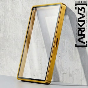 May include: A rectangular, gold-colored frame with a clear glass panel. The frame has a sleek, modern design and is leaning against a textured, gray wall. The text "ARKIV3" is visible on the right side of the frame, along with additional text.