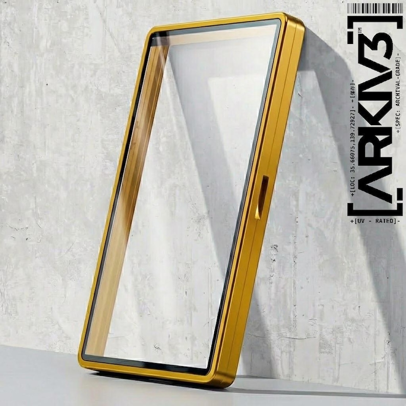 May include: A rectangular, gold-colored frame with a clear glass panel. The frame has a sleek, modern design and is leaning against a textured, gray wall. The text "ARKIV3" is visible on the right side of the frame, along with additional text.