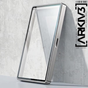 May include: A rectangular, silver-framed display case with a clear glass front, leaning against a textured, light gray wall. The case has a sleek, modern design. The text "ARKIV3" and other technical details are printed on the right side.