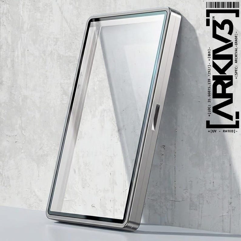 May include: A rectangular, silver-framed display case with a clear glass front, leaning against a textured, light gray wall. The case has a sleek, modern design. The text "ARKIV3" and other technical details are printed on the right side.