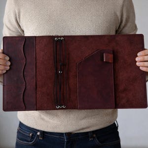Puede incluir: Funda de diario de cuero burdeos abierta, con múltiples bolsillos y un portalápices. La cubierta se mantiene abierta, mostrando el diseño interior y el cierre elástico. El cuero tiene un aspecto texturizado.