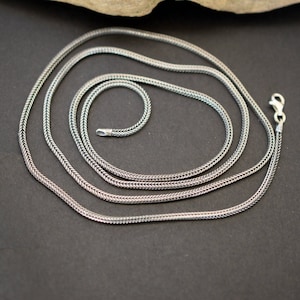 May include: A silver chain necklace with a delicate, woven design. The chain is layered on a dark background.