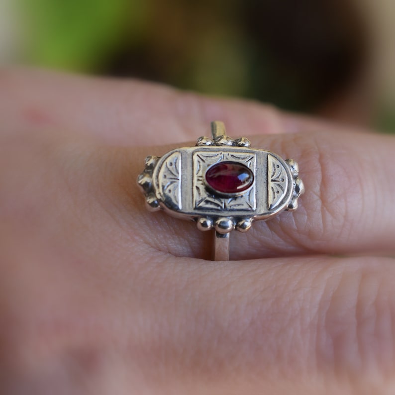 Pu&ograve; includere: Un anello d'argento con una pietra preziosa rossa ovale incastonata in un design dettagliato e ornato.