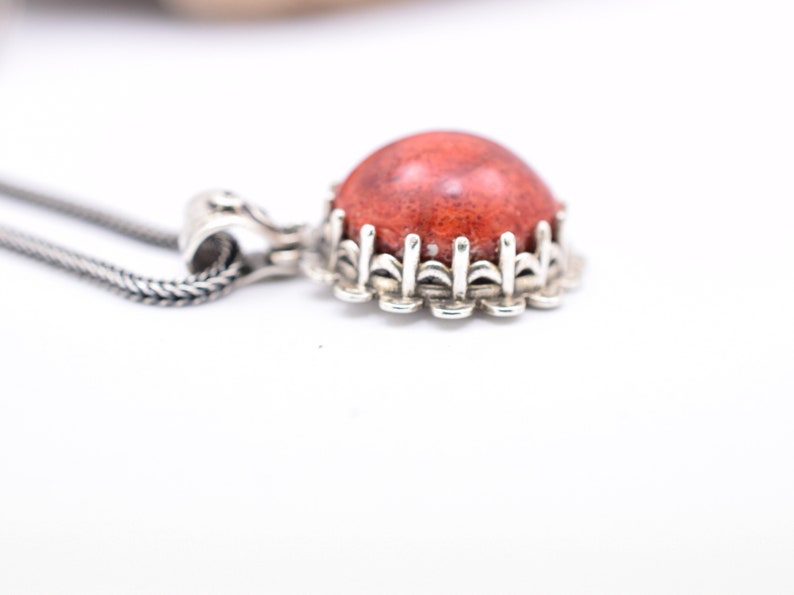 May include: A silver necklace with a red coral pendant. The pendant is set in a silver bezel with a decorative border.