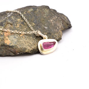 Könnte beinhalten: Eine silberne Halskette mit einem Anhänger, der einen leuchtend rosa Edelstein zeigt. Der Anhänger hat eine einzigartige, unregelmäßige Form und ist in einer goldfarbenen Lünette gefasst. Die Kette ist filigran und die Halskette wird vor einem dunklen, strukturierten Hintergrund präsentiert.