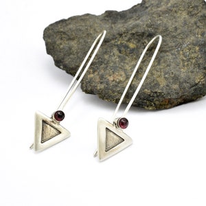May include: A pair of silver dangle earrings with a triangle-shaped pendant and a small red gemstone at the top of each pendant.