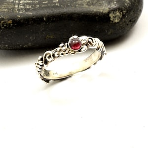 Puede incluir: Un anillo de plata con un diseño floral detallado, con una pequeña gema roja y redonda. La banda del anillo está adornada con hojas y pequeñas decoraciones en forma de cuentas. El anillo se presenta sobre un fondo blanco con una piedra gris oscura.
