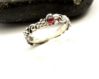 May include: A silver ring with a detailed floral design, featuring a small, round, red gemstone. The ring's band is adorned with leaves and small, bead-like embellishments. The ring is set against a white background with a dark gray stone.