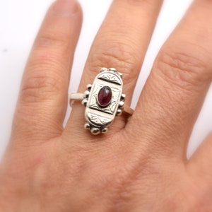 Pu&ograve; includere: Un anello d'argento con una pietra preziosa di forma ovale di colore rosso scuro incastonata in un design inciso dettagliato.