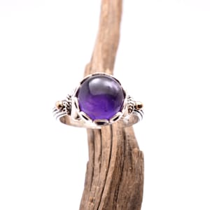 Op de afbeelding: Een zilveren ring met een grote, ronde, dieppaarse amethist edelsteen. De ring heeft sierlijke details met gouden accenten. De ring wordt gepresenteerd op een stuk drijfhout tegen een witte achtergrond.