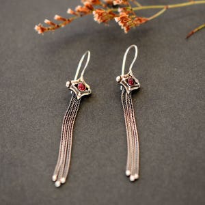 Può includere: Un paio di orecchini pendenti in argento con accenti di pietre preziose rosse. Gli orecchini presentano un design delicato a catena e un piccolo ciondolo di forma quadrata con una pietra preziosa rossa al centro.