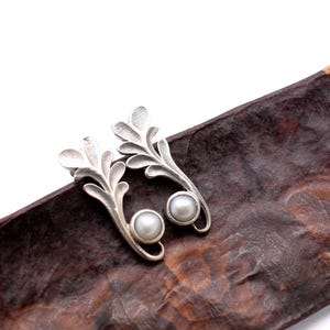 Puede incluir: Un par de pendientes plateados con forma de hoja, cada uno con una perla en la base. Los pendientes se exhiben sobre una superficie texturizada de color marrón oscuro. Diseño inspirado en la naturaleza.