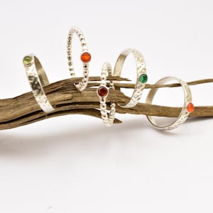 Puede incluir: Cinco anillos de plata con piedras preciosas de colores, incluyendo verde, rojo y naranja, se exhiben sobre un trozo de madera flotante. Los anillos tienen varios diseños, incluyendo bandas texturizadas y con cuentas. El fondo es blanco.