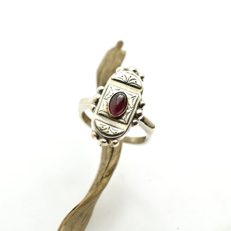 Pu&ograve; includere: Un anello d'argento con una pietra preziosa rossa ovale incastonata in un design dettagliato e ornato.