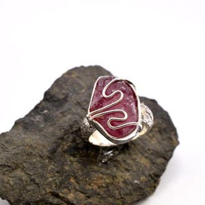 Peut inclure: Bague en argent avec une grosse pierre rouge foncé irrégulière. La pierre est sertie dans un motif de fil d'argent. La bague repose sur une roche gris foncé sur fond blanc. Le bracelet de la bague a un motif texturé.