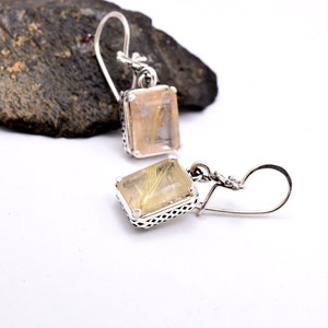 Pode incluir: Um par de brincos de prata com uma pedra de quartzo retangular transparente com inclusões douradas. Os brincos têm um design delicado e vazado.