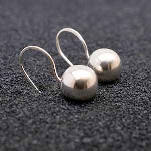 May include: A pair of silver ball earrings with curved ear wires. The earrings have a polished, reflective surface and are set against a dark, textured background. The earrings are a simple, elegant design.