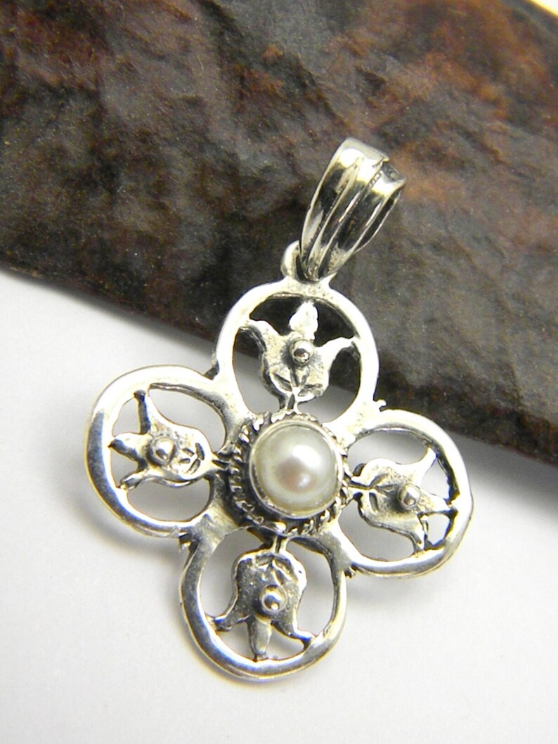 May include: A silver pendant with a delicate, intricate design featuring four stylised flowers surrounding a white pearl in the centre. The pendant has a loop at the top for attaching to a chain.