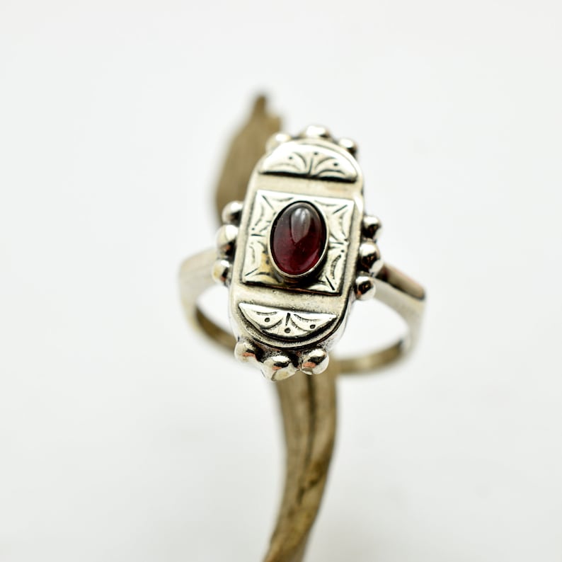 Pu&ograve; includere: Un anello d'argento con una pietra preziosa di granato di forma ovale incastonata in una cornice decorativa. L'anello presenta intricati motivi floreali e una finitura testurizzata.