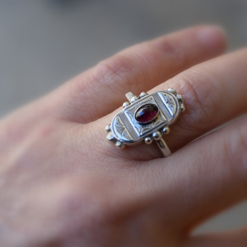 Pu&ograve; includere: Un anello d'argento con una pietra preziosa rossa ovale incastonata in un design inciso dettagliato.