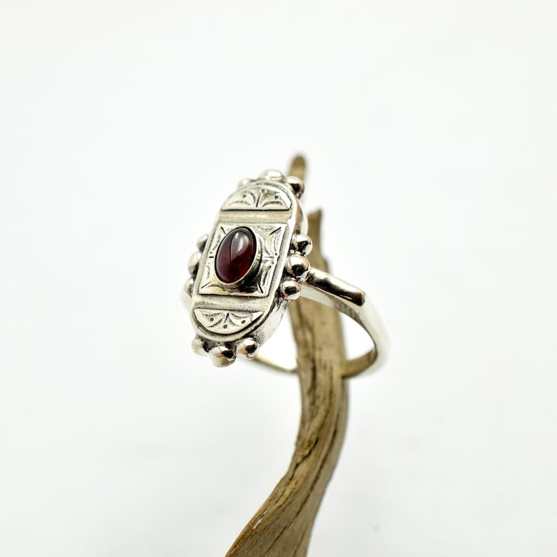 Pu&ograve; includere: Un anello d'argento con una montatura ovale che presenta una pietra preziosa rossa e dettagli incisi intricati.