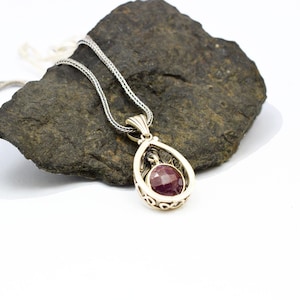 May include: A silver pendant necklace featuring a round, faceted deep red gemstone set within an ornate, teardrop-shaped silver frame with intricate scrollwork details. The pendant hangs from a delicate silver chain, showcasing a polished finish.