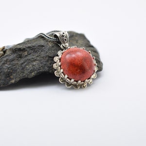 May include: A silver pendant with a red coral stone set in a decorative silver frame. The pendant is on a black chain and is resting on a gray rock.