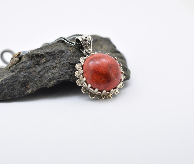 May include: A silver pendant with a red coral stone set in a decorative silver frame. The pendant is on a black chain and is resting on a gray rock.