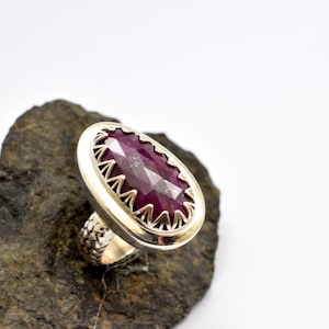 May include: A silver ring with a large, oval-shaped, faceted ruby gemstone. The ruby is a deep red color and is set in a silver bezel with a decorative edge. The ring is displayed on a dark gray rock.