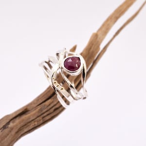 Op de afbeelding: Een zilveren ring met een grote, ovaalvormige, dieprode edelsteen. De ring heeft een uniek, open ontwerp met meerdere banden en kleine gouden accenten. De ring wordt gepresenteerd op een stuk drijfhout tegen een witte achtergrond.