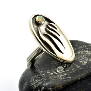 Può includere: Un anello d'argento con un design ovale che presenta una mano stilizzata e una piccola pietra di opale.