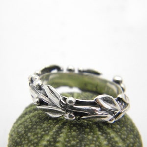 Könnte beinhalten: Ein silberner Ring mit einem filigranen Design aus Blättern und Beeren. Der Ring liegt auf einer grünen, strukturierten Oberfläche.