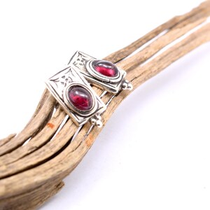 May include: Silver-toned rectangular earrings with a red gemstone center. The earrings have decorative detailing and are displayed on a piece of driftwood. The gemstones are oval-shaped and set in a silver frame.