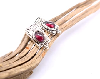 May include: Silver-toned rectangular earrings with a red gemstone center. The earrings have decorative detailing and are displayed on a piece of driftwood. The gemstones are oval-shaped and set in a silver frame.