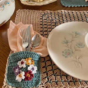 Puede incluir: Un porta servilletas de crochet color turquesa con flores de colores, una servilleta de melocotón, un tenedor, un cuchillo y una cuchara sobre un mantel individual de crochet beige. Un plato blanco con un diseño floral está sobre la mesa.