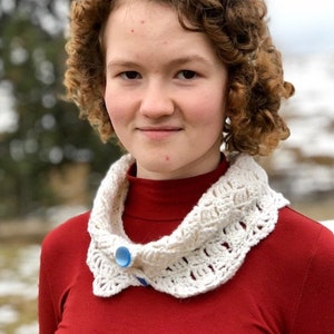 May include: A young person with curly brown hair wearing a red turtleneck sweater with a white crocheted collar and a blue button.