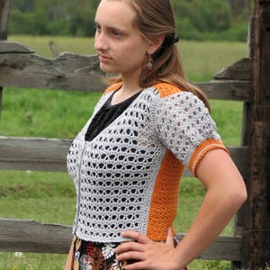 May include: A woman wearing a crocheted cardigan with a white and orange color scheme. The cardigan has a lacy pattern and a short sleeve. The woman is wearing a floral skirt and has her hair pulled back.
