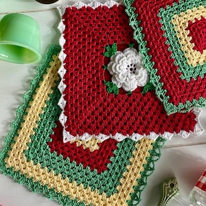 Pode incluir: Pegadores de panela de crochê feitos à mão em vermelho, verde, branco e amarelo. Os pegadores de panela quadrados apresentam um design floral e bordas onduladas. Os pegadores de panela são dispostos sobre uma superfície de madeira branca, com utensílios de cozinha ao fundo.