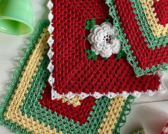 May include: Handmade crochet potholders in red, green, white, and yellow. The square potholders feature a floral design and scalloped edges. The potholders are arranged on a white wooden surface, with kitchen utensils in the background.